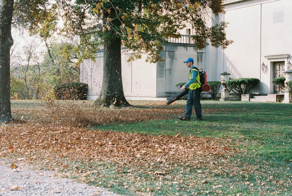 A landscaper using a leaf blower to clear fallen leaves from a lawn in front of a large house.