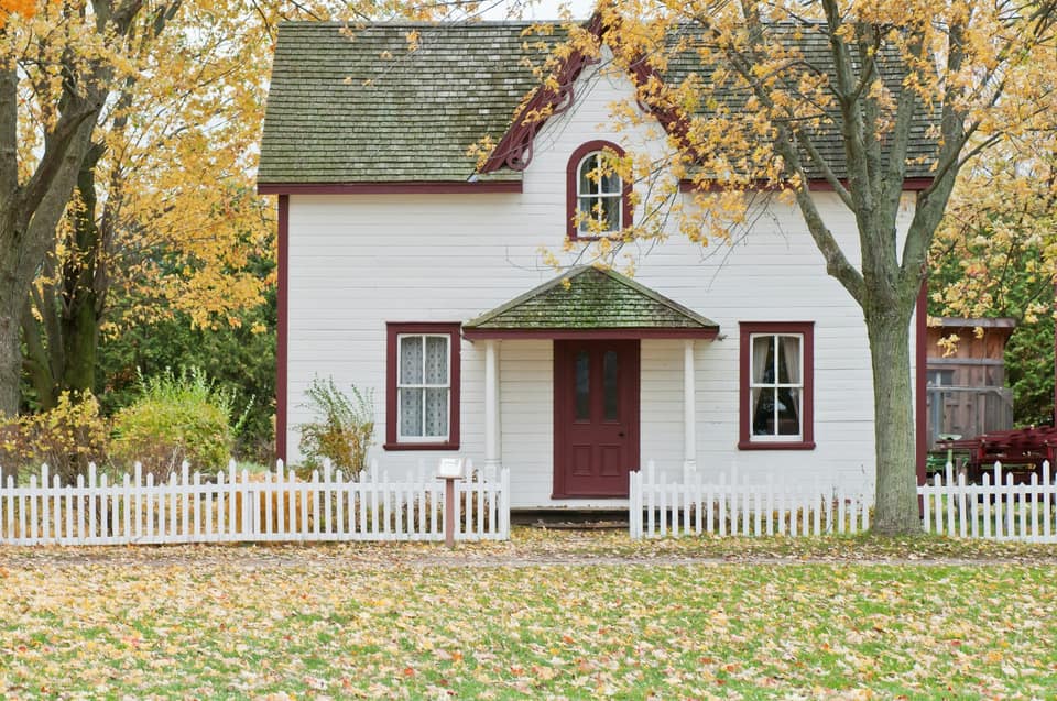 A white farmhouse with a picket fence under maple trees in full orange and gold fall color.